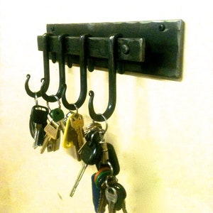 May include: A black metal key rack with four hooks, mounted on a wall. A set of keys is hanging on the hooks.