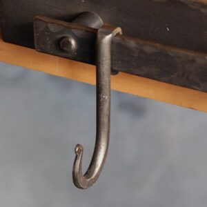 Op de afbeelding: Een zwarte metalen haak met een gebogen uiteinde, bevestigd aan een zwarte metalen balk.