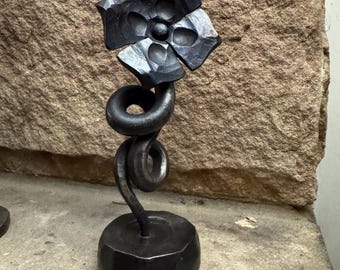 May include: A dark gray metal sculpture of a flower with four hammered petals and a central circular bud. The stem is formed by two distinct, spiraling metal coils that rise from a heavy, cylindrical, faceted metal base with a dark finish.