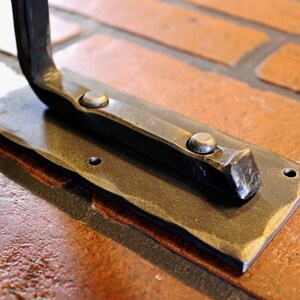 May include: Close-up of a hand-forged metal bracket. The bracket has a rectangular base with a hammered edge and two rivets. The curved arm extends from the base. The metal has a dark, aged finish. The bracket is mounted on a brick surface.