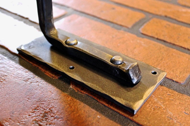 May include: Close-up of a hand-forged metal bracket. The bracket has a rectangular base with a hammered edge and two rivets. The curved arm extends from the base. The metal has a dark, aged finish. The bracket is mounted on a brick surface.