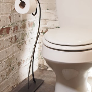 May include: A black metal toilet paper holder with a white roll of toilet paper. The holder has a decorative scroll design and stands on a rectangular base. The holder is in a bathroom with a white toilet and white brick walls.