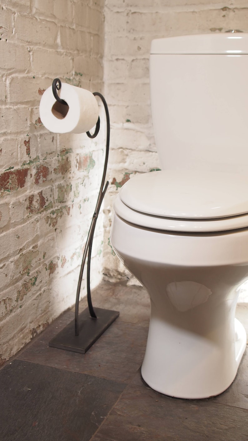 May include: A black metal toilet paper holder with a white roll of toilet paper. The holder has a decorative scroll design and stands on a rectangular base. The holder is in a bathroom with a white toilet and white brick walls.