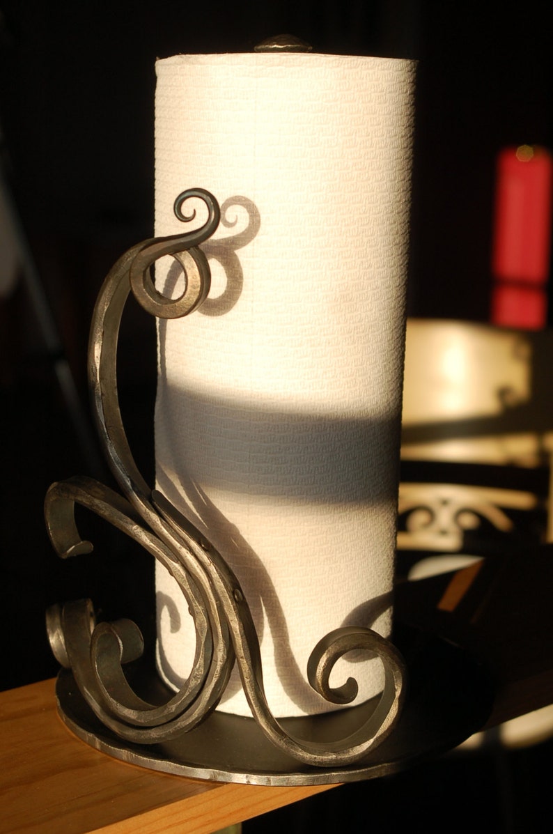 May include: A black metal paper towel holder with a decorative scroll design. The holder is holding a roll of white paper towels.