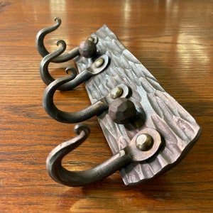 May include: A rustic metal coat rack with three hooks and a hammered wood base. The hooks are hand-forged with decorative swirls and the base has a textured surface.