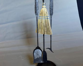 May include: A black wrought iron fireplace tool set featuring a stand with a circular base, a shovel, a poker, and a broom with natural tan bristles and blue thread accents. The tools have textured, hammered handles and hang from a curved top bracket.