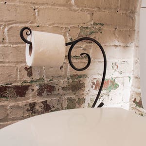 May include: A white toilet paper roll is mounted on a black metal toilet paper holder with a decorative scroll design. The holder is attached to a white toilet bowl. The toilet bowl is in front of a brick wall with white paint and green and brown paint chips.