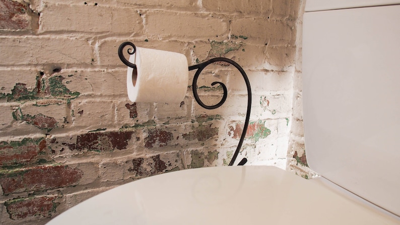 May include: A white toilet paper roll is mounted on a black metal toilet paper holder with a decorative scroll design. The holder is attached to a white toilet bowl. The toilet bowl is in front of a brick wall with white paint and green and brown paint chips.