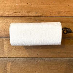 May include: A roll of white paper towels on a decorative metal holder with a leaf design. The holder is mounted on a wooden surface with horizontal planks. The paper towels are textured and ready for use.