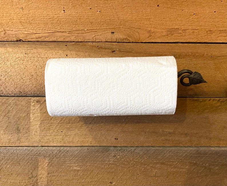 May include: A roll of white paper towels on a decorative metal holder with a leaf design. The holder is mounted on a wooden surface with horizontal planks. The paper towels are textured and ready for use.