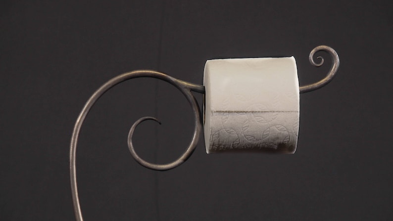 May include: A metal toilet paper holder with a decorative scroll design. The holder is made of dark metal and has a single roll of toilet paper on it.