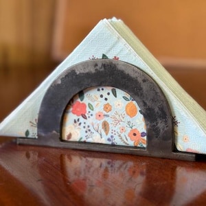 May include: A metal napkin holder with a semi-circular design, holding a stack of floral-patterned napkins. The napkins have a light blue background with orange and red flowers. The holder is made of dark metal.
