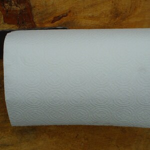 May include: A black metal paper towel holder with a decorative scroll design. The holder is mounted to a wooden surface and has a white paper towel roll attached.