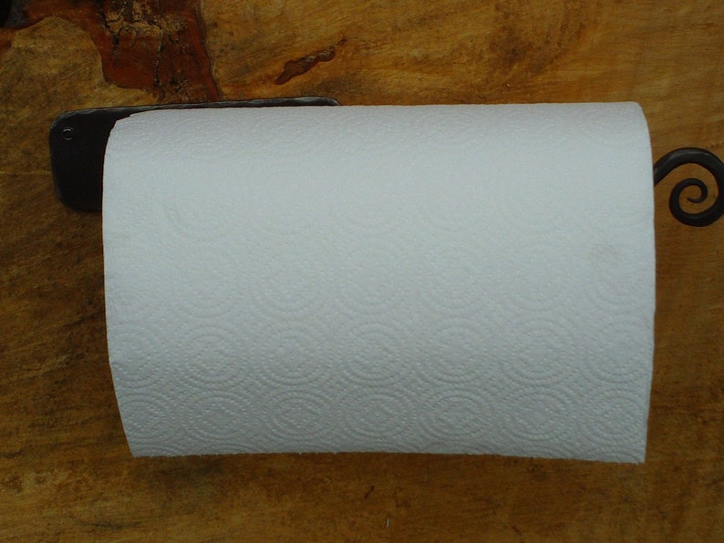 May include: A black metal paper towel holder with a decorative scroll design. The holder is mounted to a wooden surface and has a white paper towel roll attached.