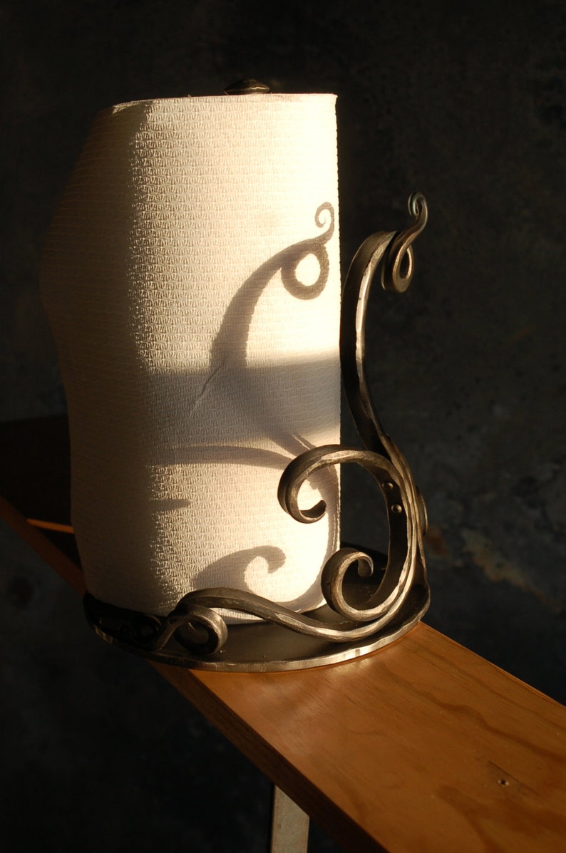 May include: A metal paper towel holder with a decorative scroll design. The holder is made of dark metal and has a white paper towel roll inside.