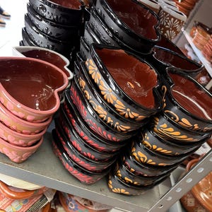 May include: Heart-shaped ceramic bowls in various colors, stacked on shelves. The bowls feature a glossy black exterior with floral designs in red and orange. Other bowls are pink and brown.