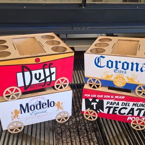 May include: Four wooden beverage carriers shaped like wagons with wheels and handles. The carriers feature logos for Duff Beer, Corona Extra, Modelo Especial, and Tecate. Each carrier has six circular cutouts for bottles and a central rectangular opening.