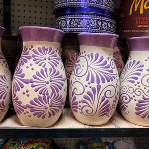 May include: A collection of handcrafted ceramic vases with a beige base and purple accents. The vases feature a detailed floral design in purple and a purple rim. The vases are displayed on a wooden shelf.