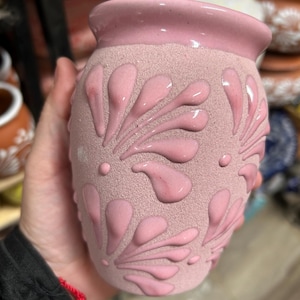 May include: A pink ceramic vase with a textured surface and raised floral designs. The vase has a slightly flared rim and is a soft, muted pink color. The raised designs are a glossy pink.