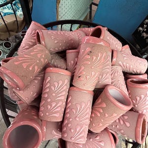 May include: A collection of pink ceramic vases with a floral design. The vases are cylindrical with a slightly flared rim and are arranged in a black metal basket. The vases have a matte finish and a glossy pink trim.