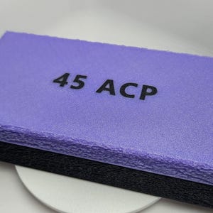 May include: A rectangular, two-tone container with a textured surface. The top is lavender, and the bottom is black. The text "45 ACP" is printed in black on the top. A small handle is visible on the side.