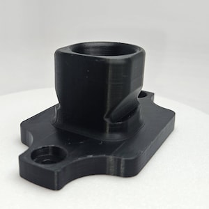 May include: A black, 3D-printed part with a square-shaped top and a rectangular base. The base has rounded corners and two circular holes. The part appears to be made of a matte plastic material.