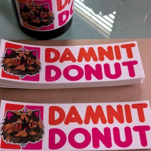 May include: Rectangular stickers featuring a graphic of a fluffy cat wearing a crown next to the text DAMNIT DONUT in orange and pink lettering. The stickers are white with a glossy finish, and a matching mug is visible in the background.
