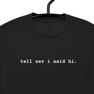 May include: Black t-shirt with the white text "tell zev i said hi." The shirt is hanging on a black hanger. The text is in a simple, sans-serif font.