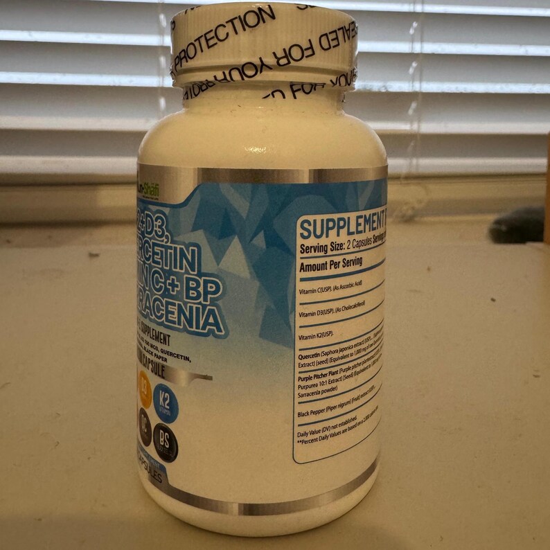 K&ouml;nnte beinhalten: Eine wei&szlig;e Erg&auml;nzungsflasche mit einem blau-silbernen Etikett. Das Etikett enth&auml;lt den Text "D3, Quercetin, Zink + BP, Placenia" und "Erg&auml;nzung". Die Flasche hat einen wei&szlig;en Deckel mit dem Text "Versiegelt f&uuml;r Ihren Schutz".