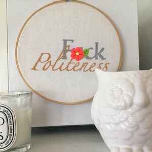 Może przedstawiać: Biała ceramiczna figurka sowy z okrągłym, białym obręczą do haftu z napisem "F*ck Politeness" w szarej i pomarańczowej nici. Obręcza do haftu wisi na białej ścianie.