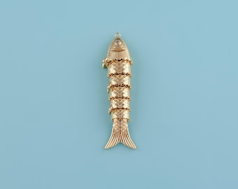 May include: Gold-colored fish pendant with a textured, scale-like design. The pendant has a loop at the top for hanging and a small, clear gem near the head. The fish's tail is split, and the background is a solid light blue.