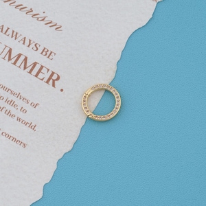 May include: A circular gold-toned hoop earring featuring a row of small, clear, sparkling stones embedded along the front surface. The jewelry piece has a hinged closure mechanism and is displayed against a textured white paper background.