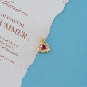 May include: A gold-toned triangular pendant featuring a central red gemstone surrounded by a border of small clear stones. The pendant includes a small circular jump ring at the top for attachment, showcasing a metallic finish and clear detailing.