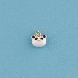May include: A small, white panda bear charm with black ears and eyes, and a gold-colored bail. The charm is set against a light blue background. This jewelry charm is a cute accessory.