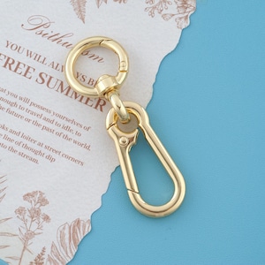 May include: A gold-toned metal swivel lobster clasp with a round ring attached to an elongated, teardrop-shaped carabiner clip. The hardware features a polished, reflective surface and a spring-loaded mechanism for secure attachment and rotation.