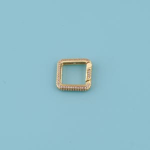 May include: A gold-colored, square-shaped clasp with a hinged side. The clasp is encrusted with small, clear gemstones around the edges. The background is a solid light blue.