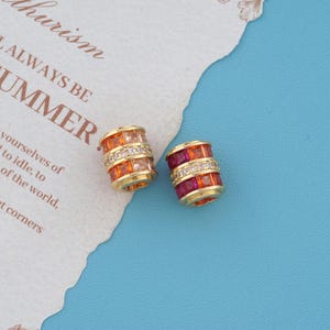 May include: Two cylindrical gold beads featuring rows of square-cut gemstones. One bead displays orange and light peach stones, while the other features deep pink and orange stones. Both beads have a central band of small, clear pavé crystals.
