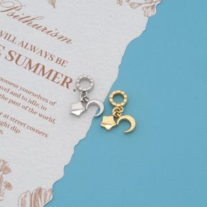 May include: Two metal charms, one silver-toned and one gold-toned, each featuring a circular ring connector attached to a small five-pointed star and a crescent moon shape. The charms are displayed against a background with printed text.