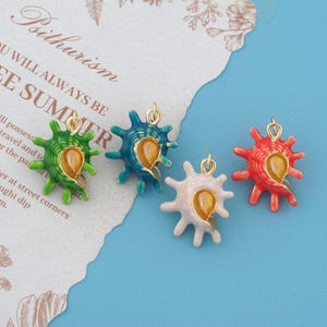 May include: Four seashell charms with gold-tone metal loops, each featuring a distinct enamel color: bright green, teal, pale pink, and vibrant red. Each charm has a gold-colored interior opening and multiple radiating points around the shell structure.