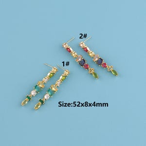 May include: Gold-toned earrings with a linear design, featuring an array of colorful gemstones. The earrings are adorned with green, blue, red, and clear stones. The text "Size: 52x8x4mm" is visible. The earrings are displayed on a light blue background.