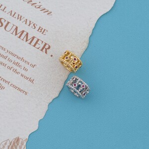 May include: Two small, cylindrical jewelry charms, one in gold-tone metal and one in silver-tone metal. Each charm features a lattice-like openwork design accented with small, round, embedded red stones arranged in a repeating pattern around the band.