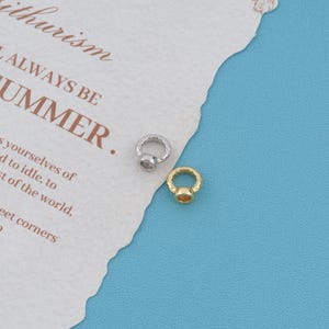 May include: Two small circular metal charms, one silver-toned and one gold-toned, each featuring a textured, patterned band and a smooth, circular center setting. The charms are positioned on a piece of paper with printed text.