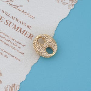 May include: A gold-toned oval charm featuring a textured surface encrusted with numerous small, clear, sparkling stones. The charm has two distinct circular cutouts in the center, creating a coffee bean-like shape with a polished metallic border.