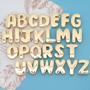 May include: A complete set of twenty-six individual gold-colored metal alphabet letters from A to Z. Each letter features a smooth, rounded, and reflective surface with a puffy, three-dimensional appearance, arranged in four horizontal rows.