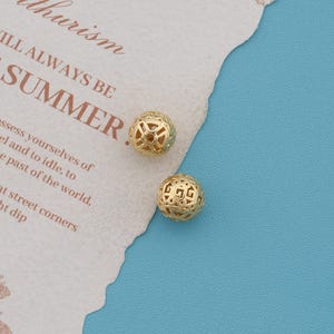 May include: Two spherical gold-toned beads featuring an intricate, openwork geometric lattice pattern. The beads are positioned on a piece of textured off-white paper with printed text that reads "thurism" and "ILL ALWAYS BE SUMMER."