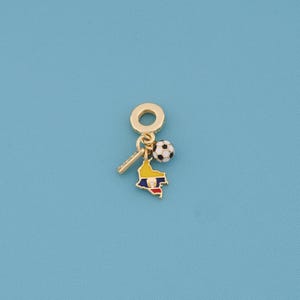 May include: A gold-colored charm with a soccer ball, a map of Colombia, and a small bar. The map is colored yellow, blue, and red. The soccer ball is black and white. The charm is on a blue background.
