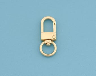 May include: A gold-colored swivel clasp with a D-shaped top and a circular ring at the bottom. The clasp is made of metal and has a shiny finish. The background is a solid light blue.