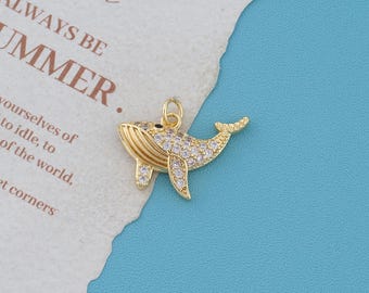 May include: A gold-toned whale charm featuring a textured body encrusted with numerous small, clear, sparkling cubic zirconia stones. The whale has a small circular jump ring attached to its top for hanging, and it is positioned on a textured paper.