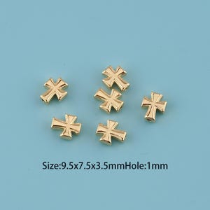 May include: Six small, gold-colored cross charms are scattered on a light blue surface. The crosses have a detailed design and a shiny finish. The text on the image indicates the size of the charms.