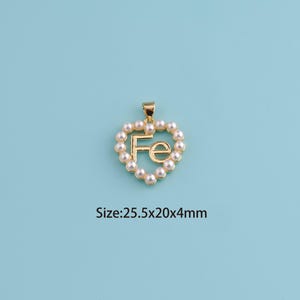 May include: A gold-colored pendant in the shape of a heart, outlined with small, round, off-white pearls. The letters "Fe" are in the center of the heart. The pendant is 25.5mm x 20mm x 4mm. The background is a solid light blue.